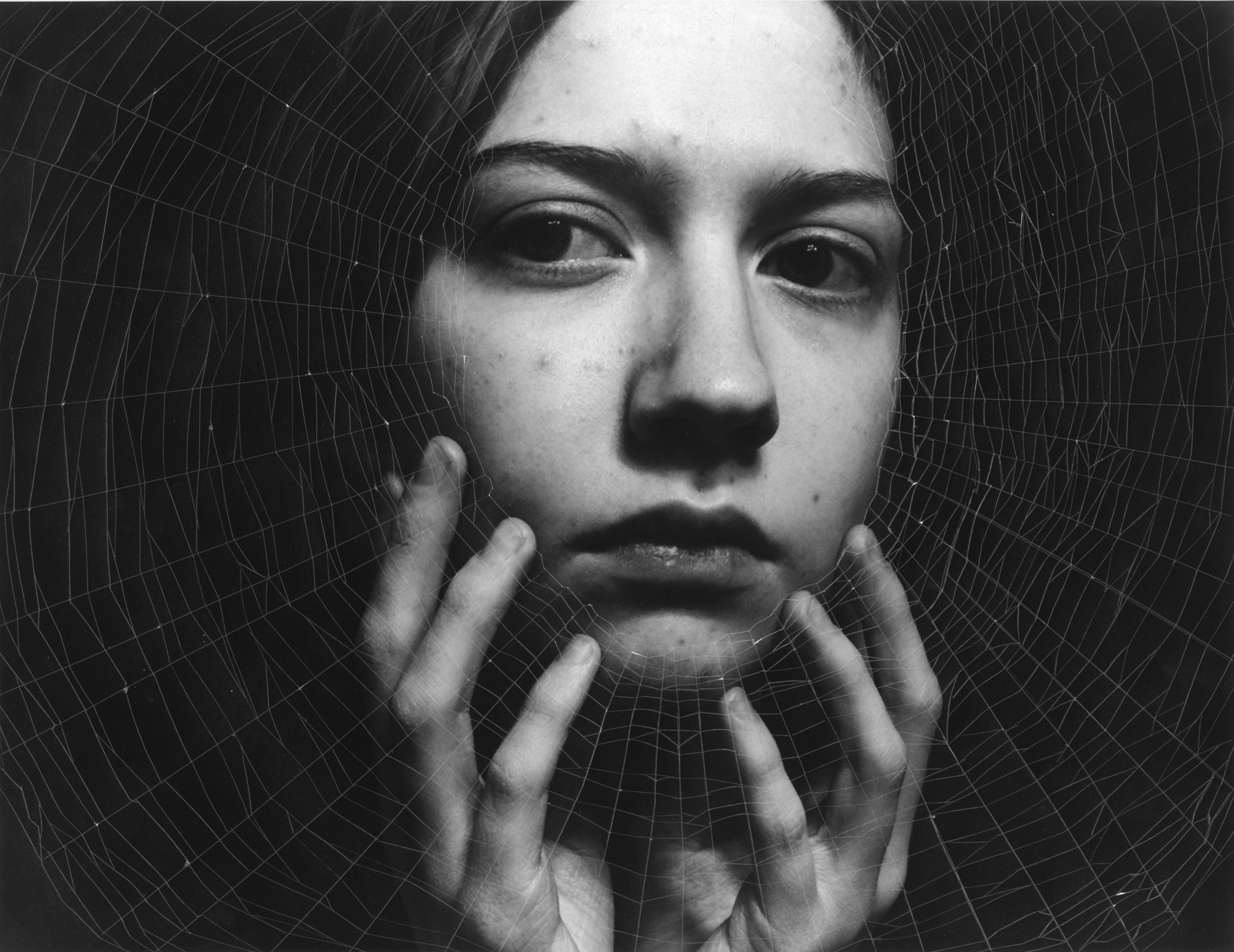 Black and white gelatin silver print of a woman looking out at the camera with her hands resting on her face, overlaid by a cutout of a spider's web.